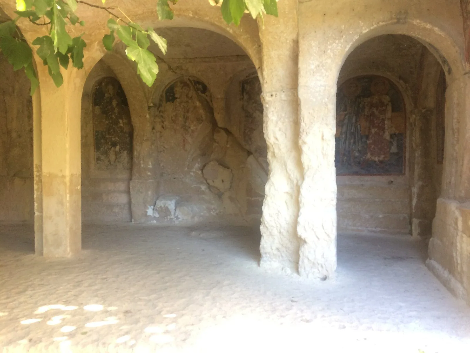 Interno di una chiesa rupestre scavata nel tufo, con archi e pilastri ricavati nella roccia e affreschi di santi nelle nicchie, illuminati dalla luce del giorno