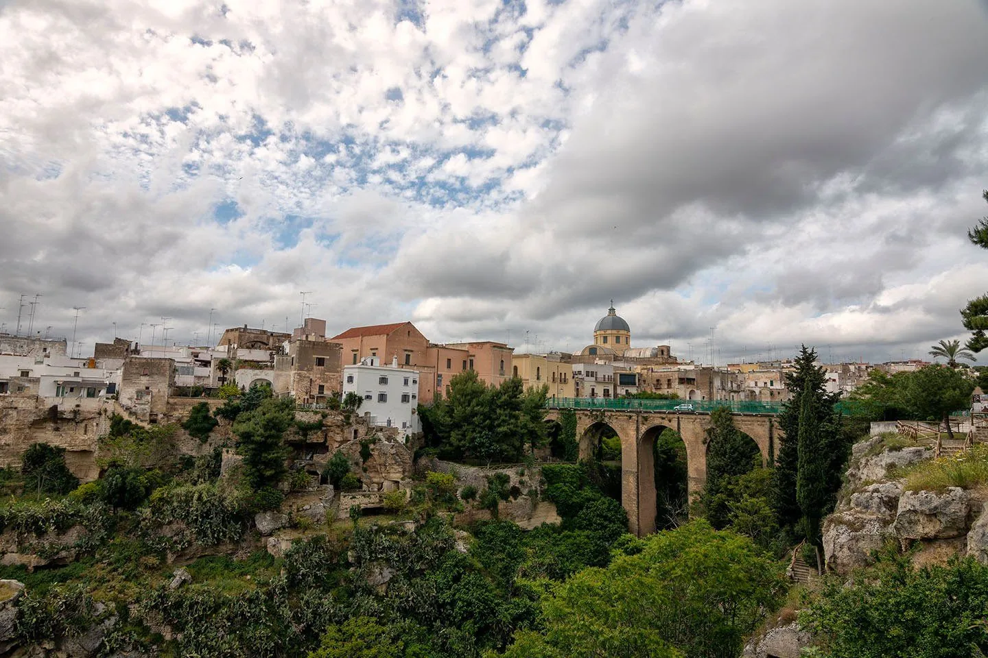 Il centro storico di Massafra affacciato sulla gravina verde, con un ponte ad arcate in pietra e la cupola del Duomo sotto un cielo nuvoloso