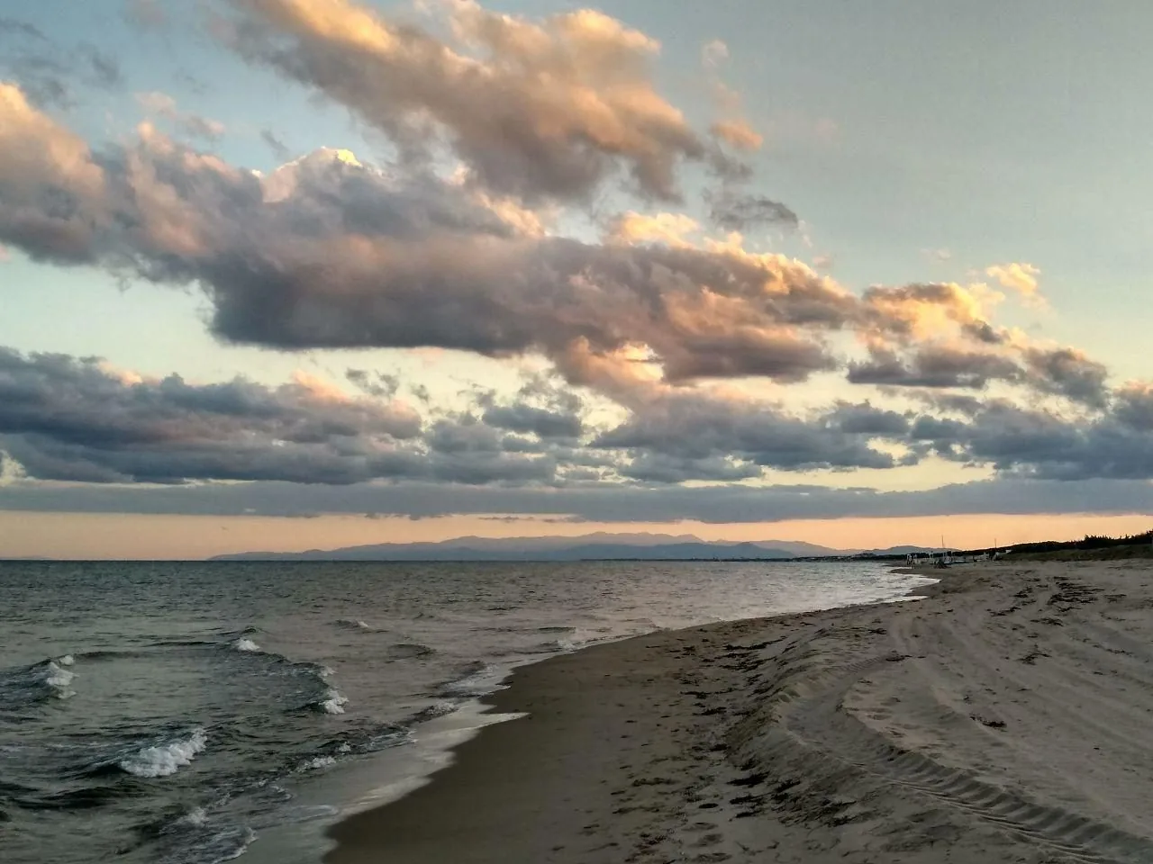 Spiaggia di sabbia a Castellaneta Marina al tramonto, con onde leggere sullo Ionio, nuvole rosate e il profilo delle montagne all'orizzonte