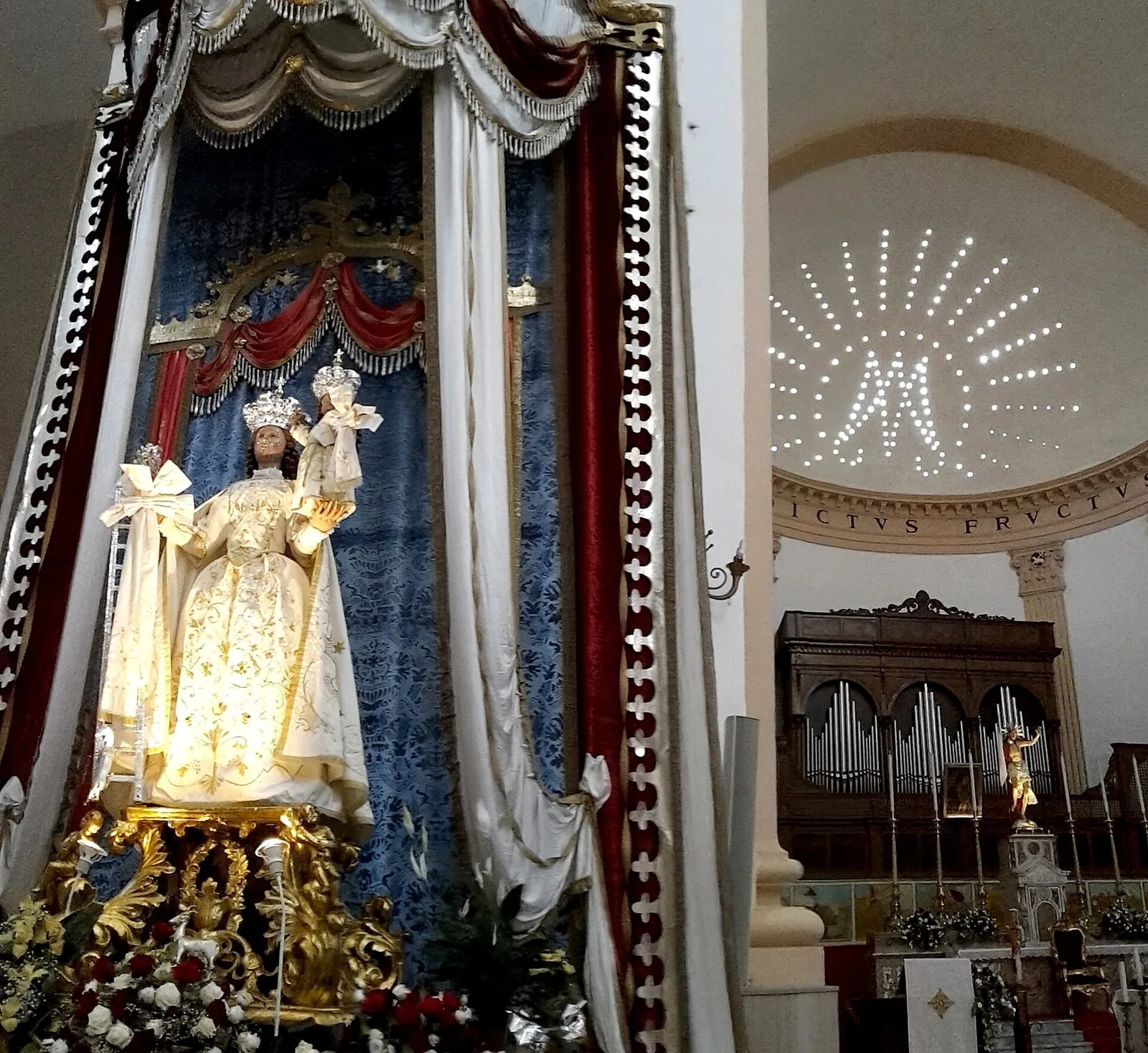 Statua della Madonna col Bambino incoronati, con abito chiaro ricamato in oro, tra drappi rossi e blu e fiori, all'interno di una chiesa con organo