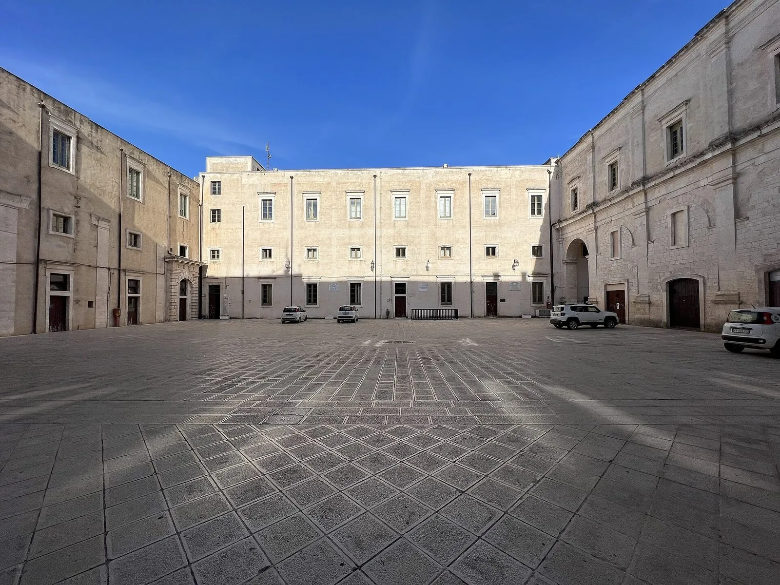 Il grande cortile lastricato del Palazzo Ducale di Martina Franca, chiuso da facciate in pietra chiara con file di finestre, sotto un cielo blu intenso