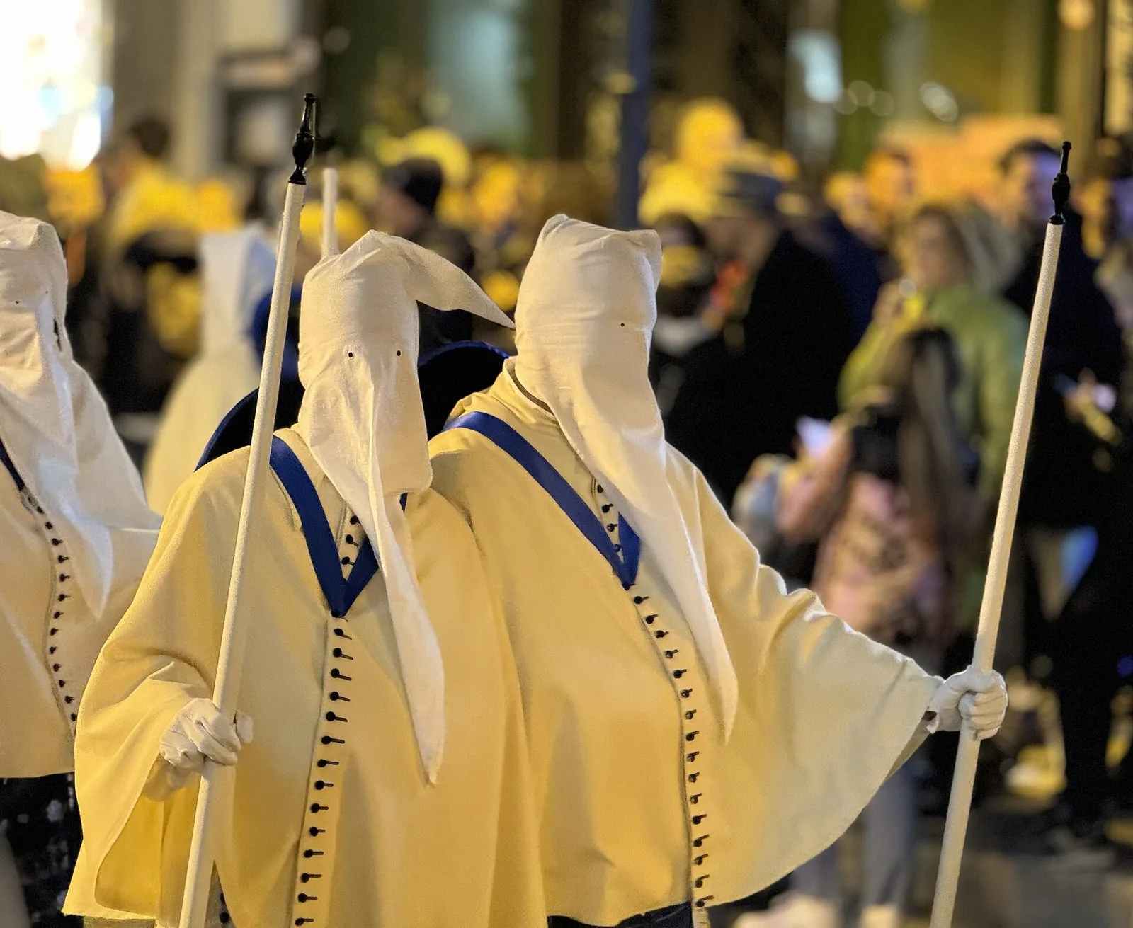 Due perdoni di Taranto con cappuccio bianco, mantella color crema bordata di blu, guanti bianchi e lungo bastone, in processione di notte tra la folla sfocata
