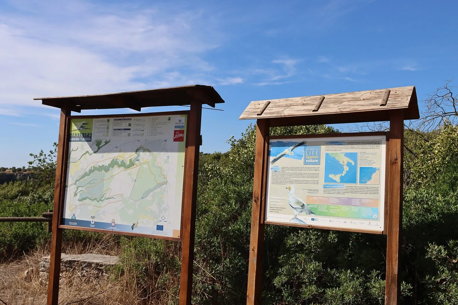 Due pannelli informativi in legno sul ciglio della Gravina di Laterza: a sinistra la mappa dei sentieri dell'oasi, a destra un pannello sul capovaccaio, tra arbusti e cielo azzurro
