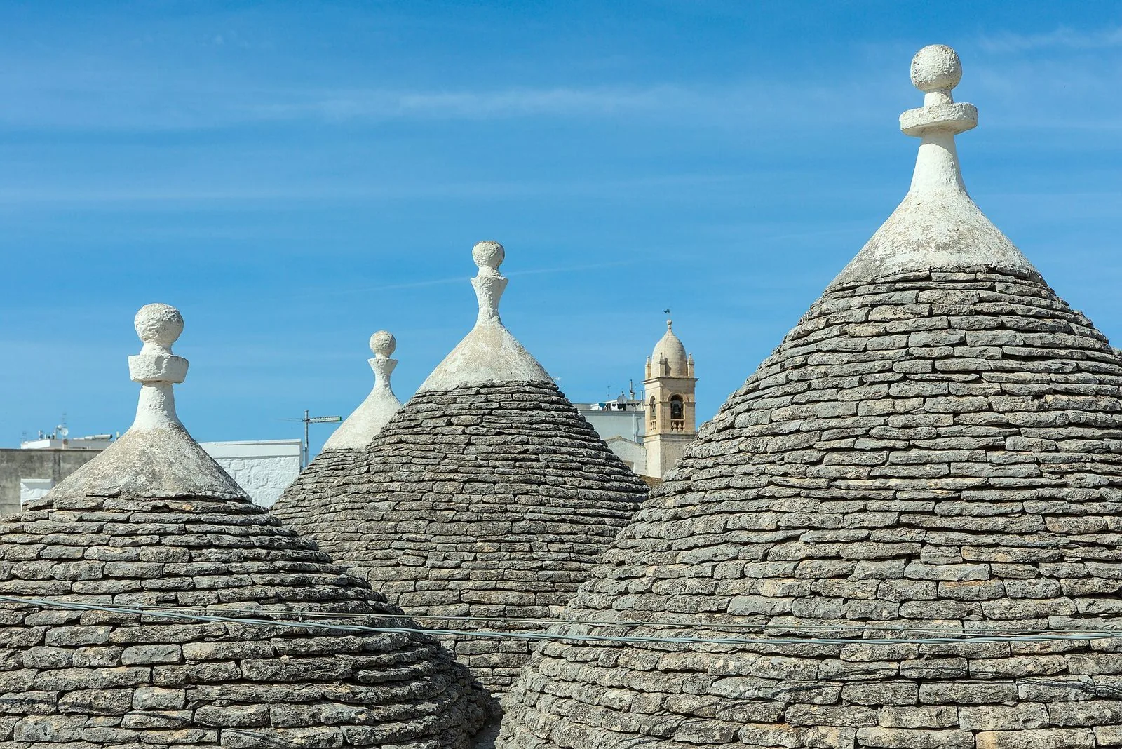 Tetti conici dei trulli di Alberobello in lastre di pietra grigia con pinnacoli bianchi, e sullo sfondo un campanile in pietra sotto il cielo azzurro