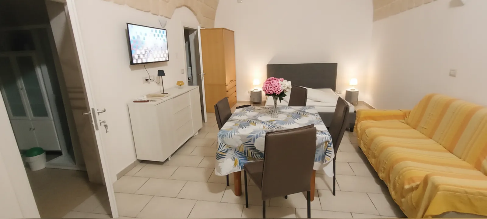 L’open space visto dall’ingresso: il mobile bianco con la TV, il tavolo apparecchiato, il letto matrimoniale e il divano letto giallo